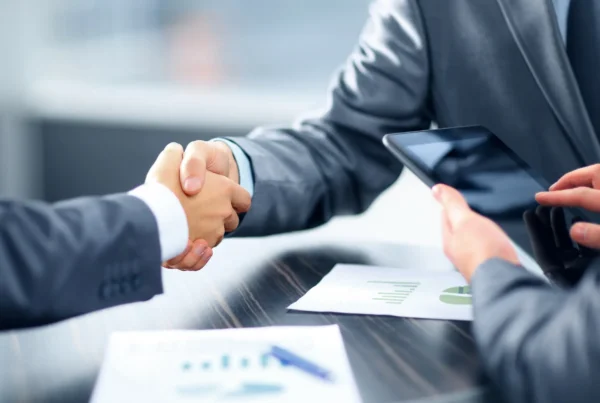 Dos ejecutivos se dan la mano en una mesa con reportes financieros mientras otra persona revisa una tableta, simbolizando cómo el historial crediticio empresarial influye en la tasa de interés.